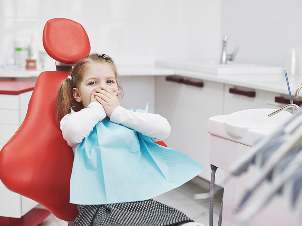 Young girl in red dental chair with hands over mouth 